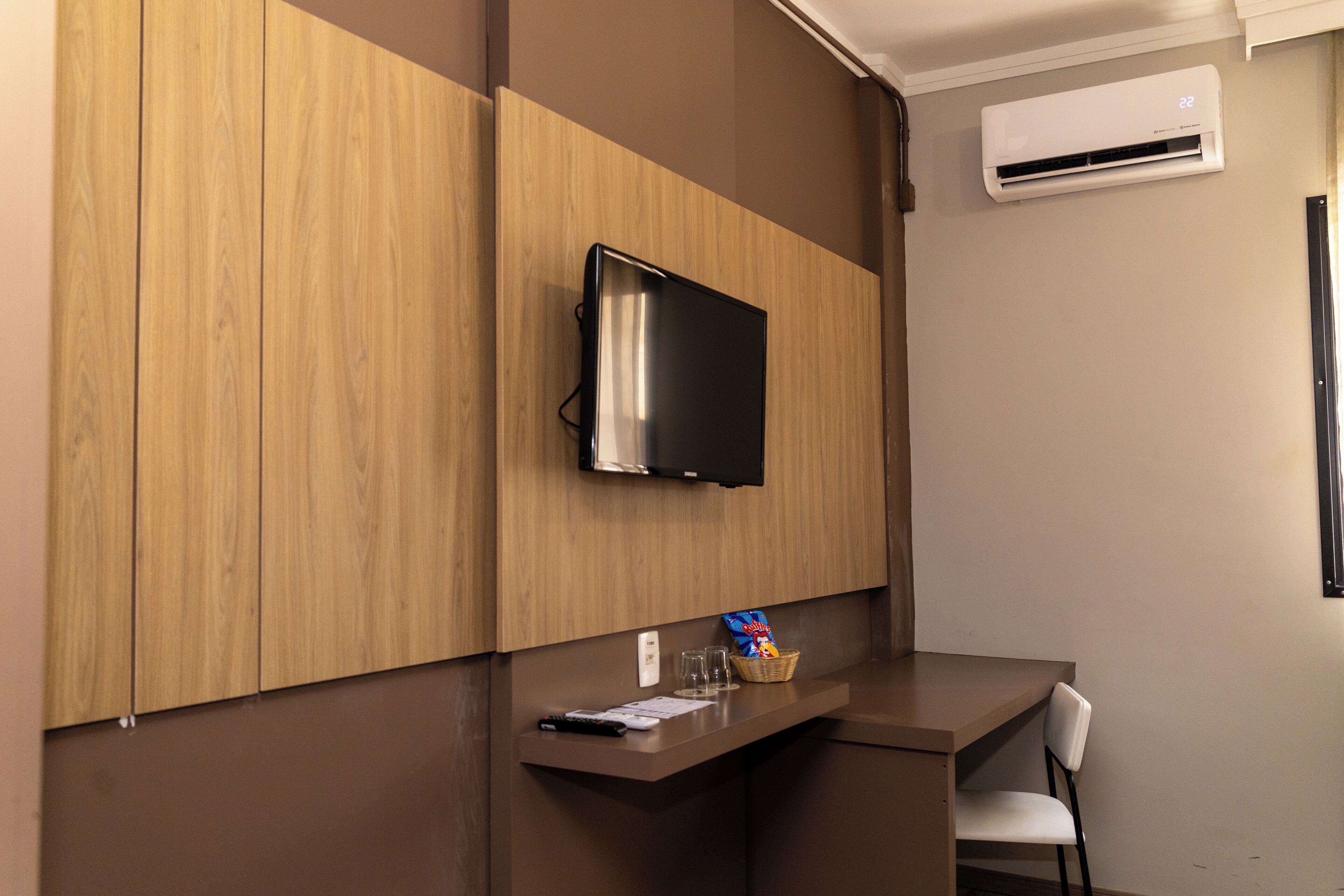 Een moderne kamer met een flatscreen televisie, bevestigd aan een houten wandpaneel, een klein aanrecht met een mandje snacks en een stoel.