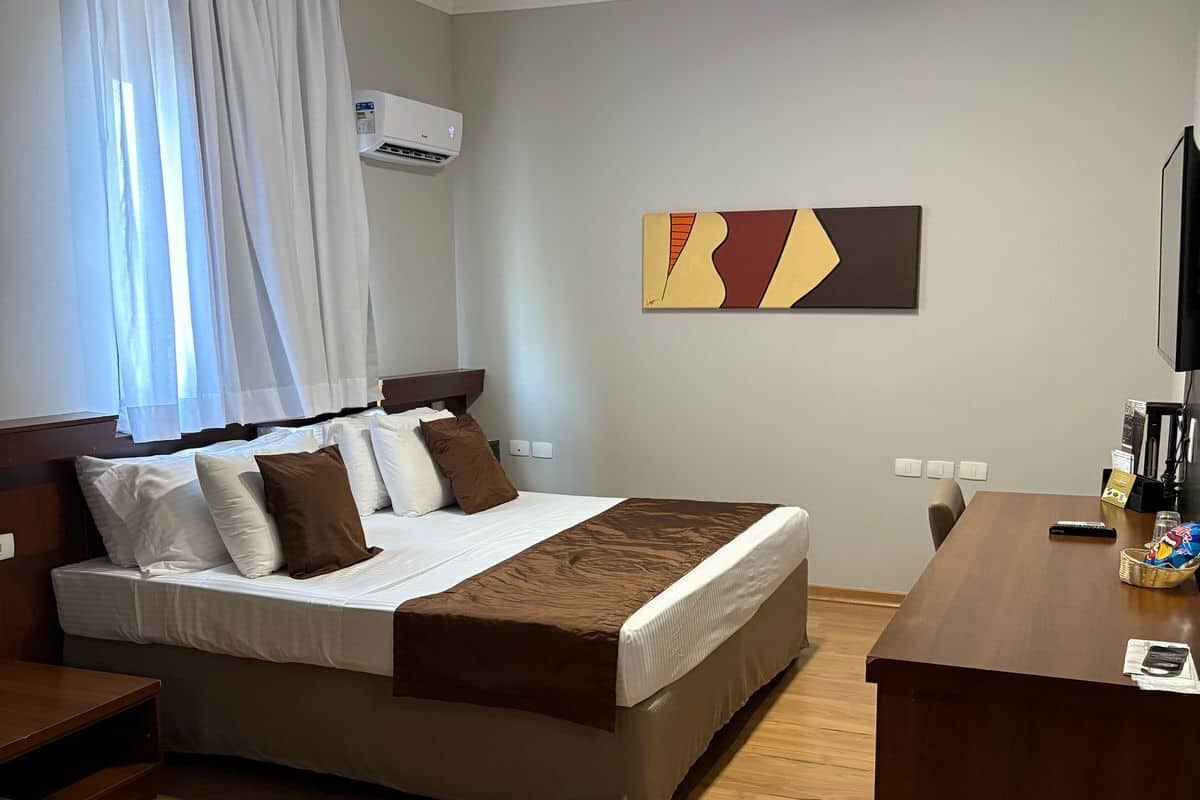 A hotel room with a bed, a desk, a chair, a TV, and an air conditioning unit.