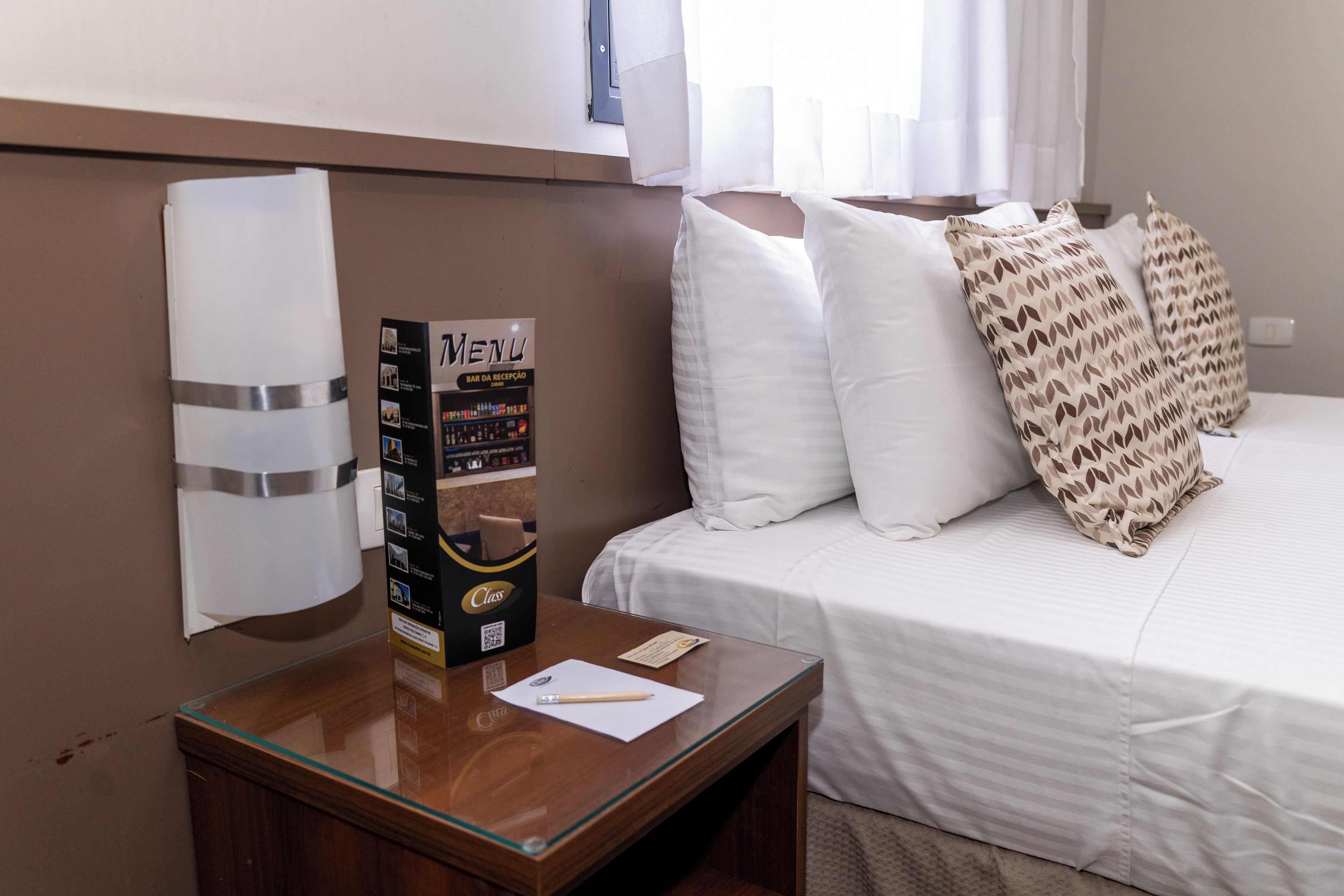 Una habitación de hotel con cama, almohadas, una mesita de noche, una mesa con cubierta de vidrio y un atril para el menú, y un aire acondicionado instalado en la pared.
