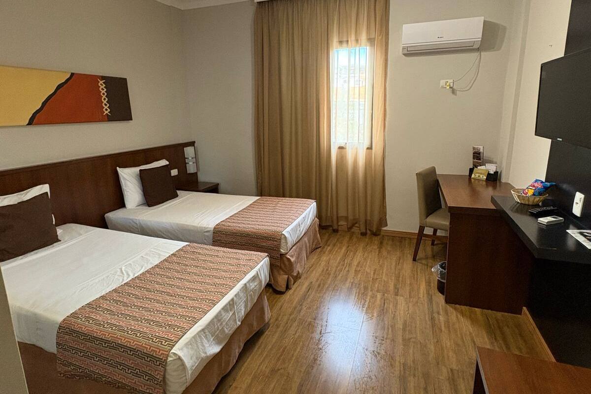 A hotel room with two beds, a desk, a chair, an air conditioner, and a TV.
