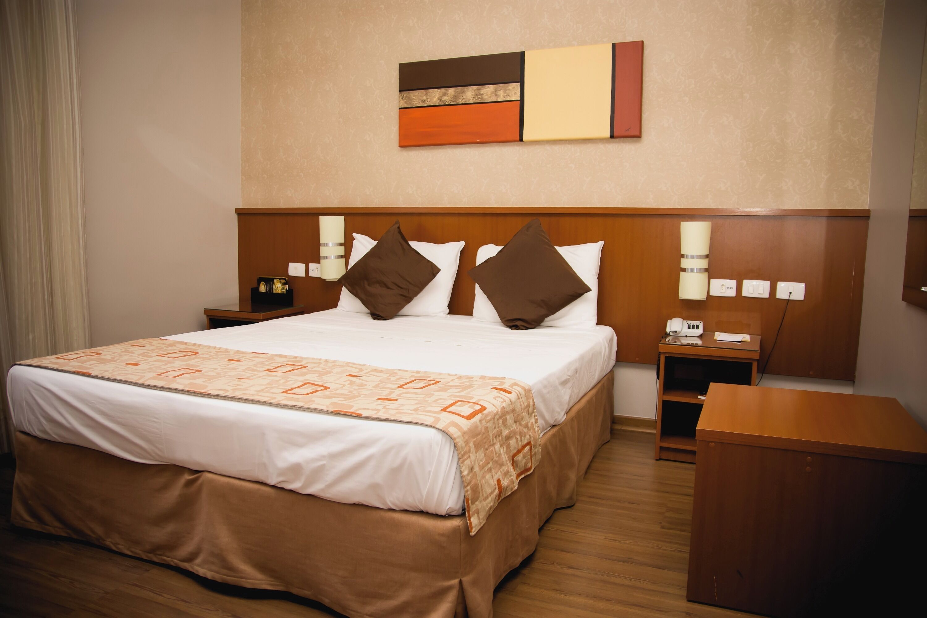 Una habitación de hotel con una cama grande, dos mesitas de noche, un escritorio de madera y una lámpara de pared.