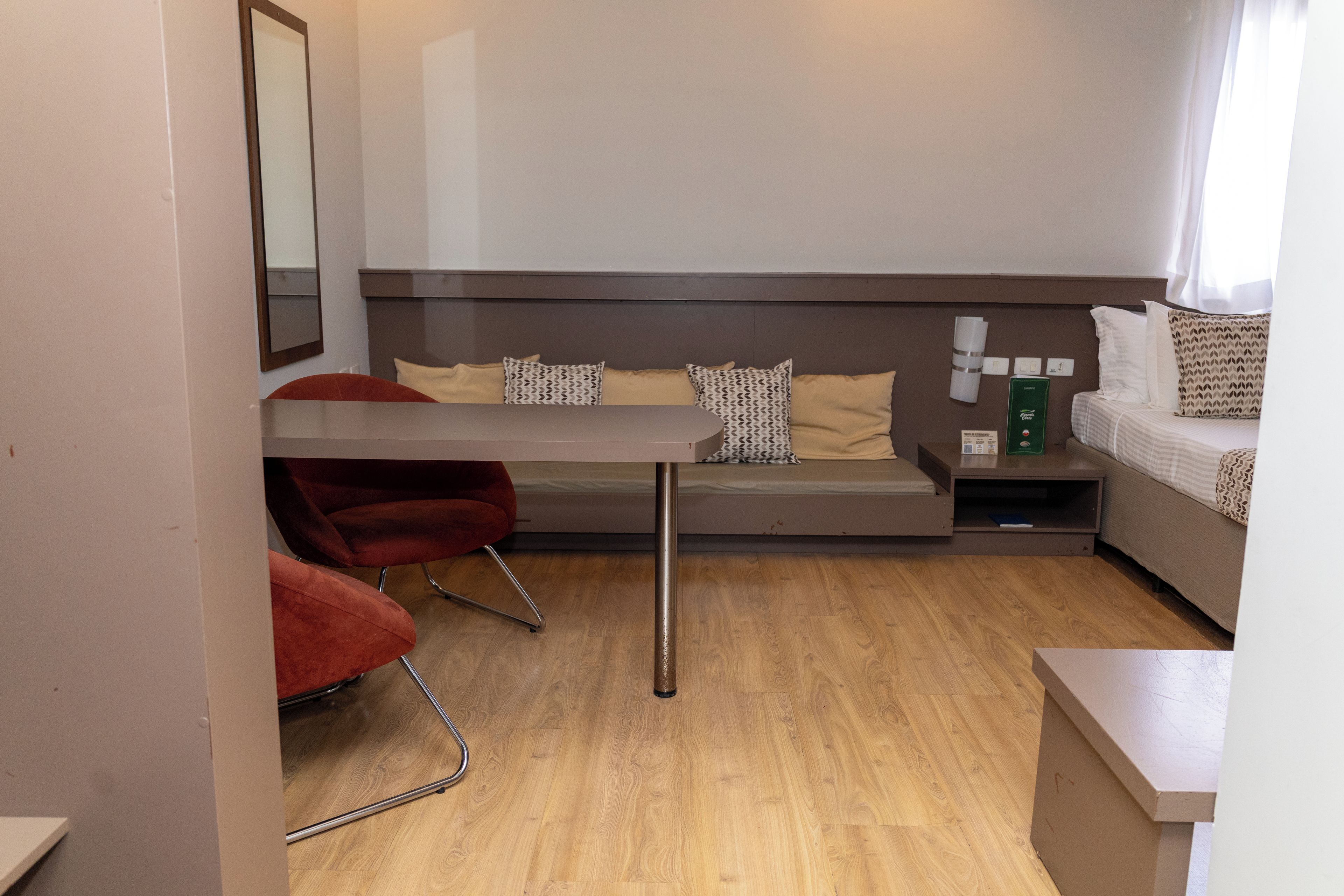 A compact hotel room with a bed, a small table, a chair, and a bedside table with a lamp.