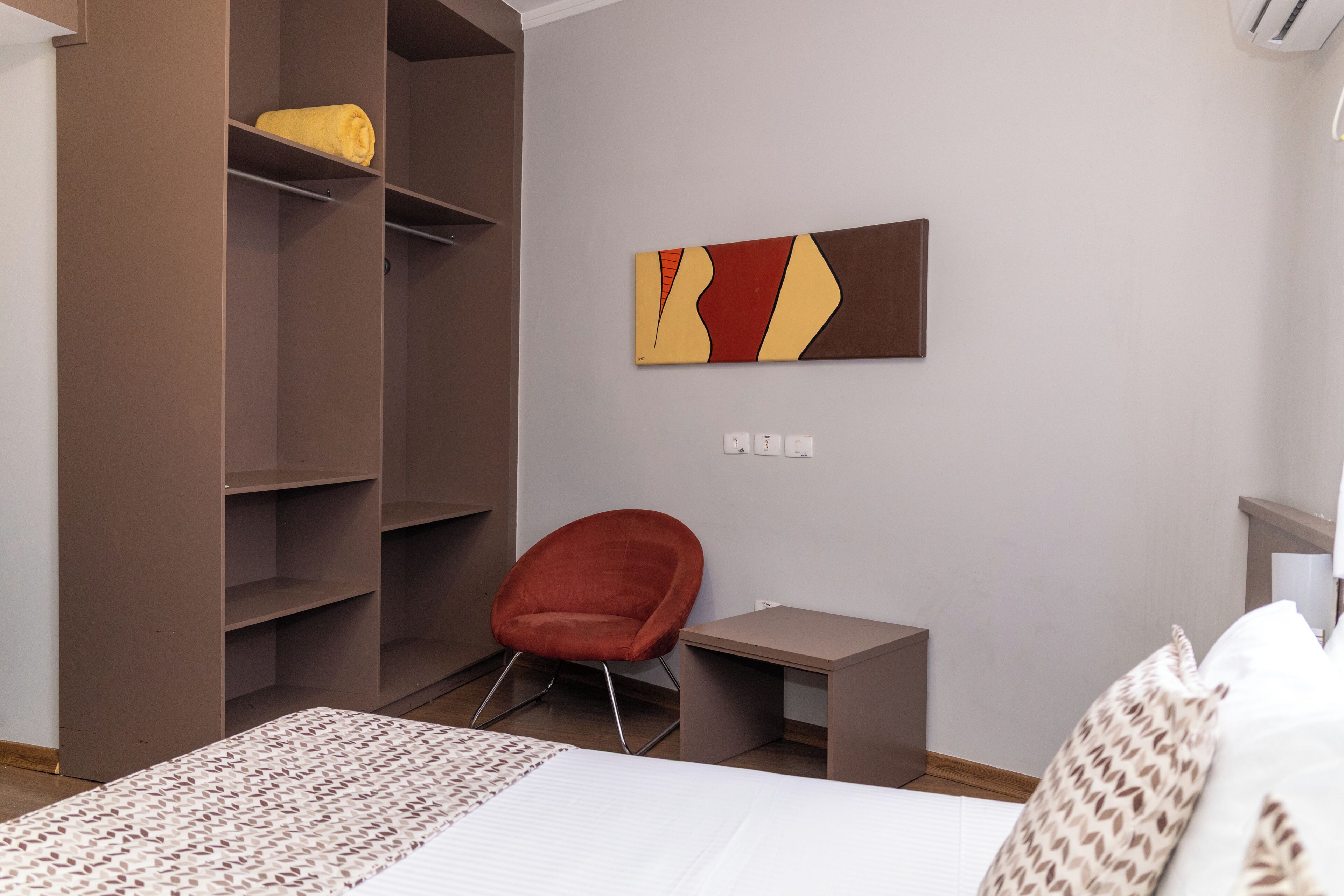 Una habitación de hotel con cama, una silla roja, una mesita, un armario y una obra de arte colgada en la pared.
