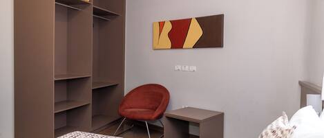 Una habitación de hotel con cama, una silla roja, una mesita, un armario y una obra de arte colgada en la pared.