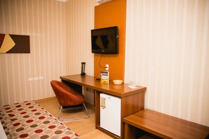 Une chambre d’hôtel avec un bureau en bois, un petit réfrigérateur, un téléviseur fixé au mur, une chaise rouge et un lit recouvert d’une couverture à pois.