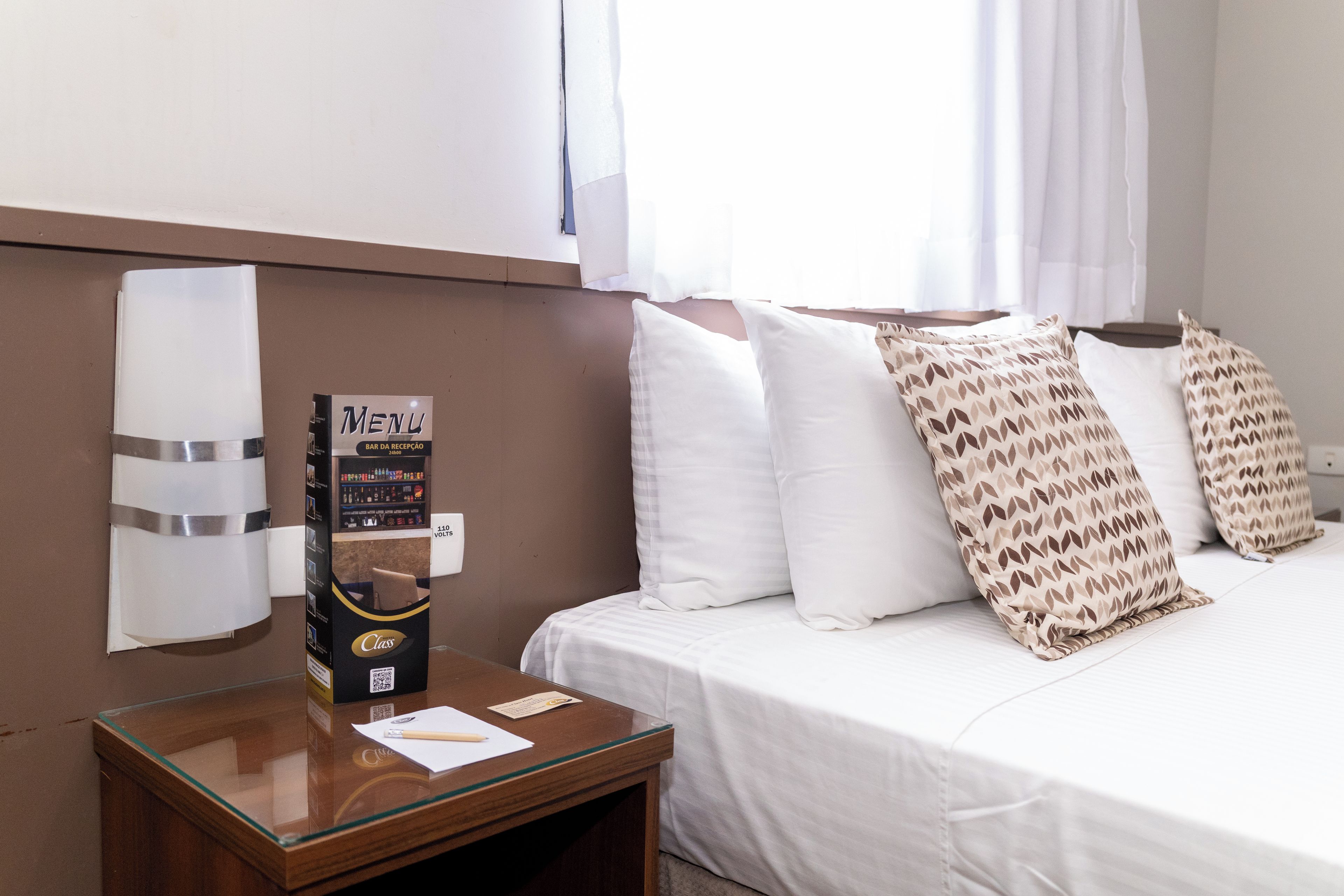 Habitación de hotel con cama, almohadas, mesita de noche y un atril para el menú, además de una ventana con cortinas.