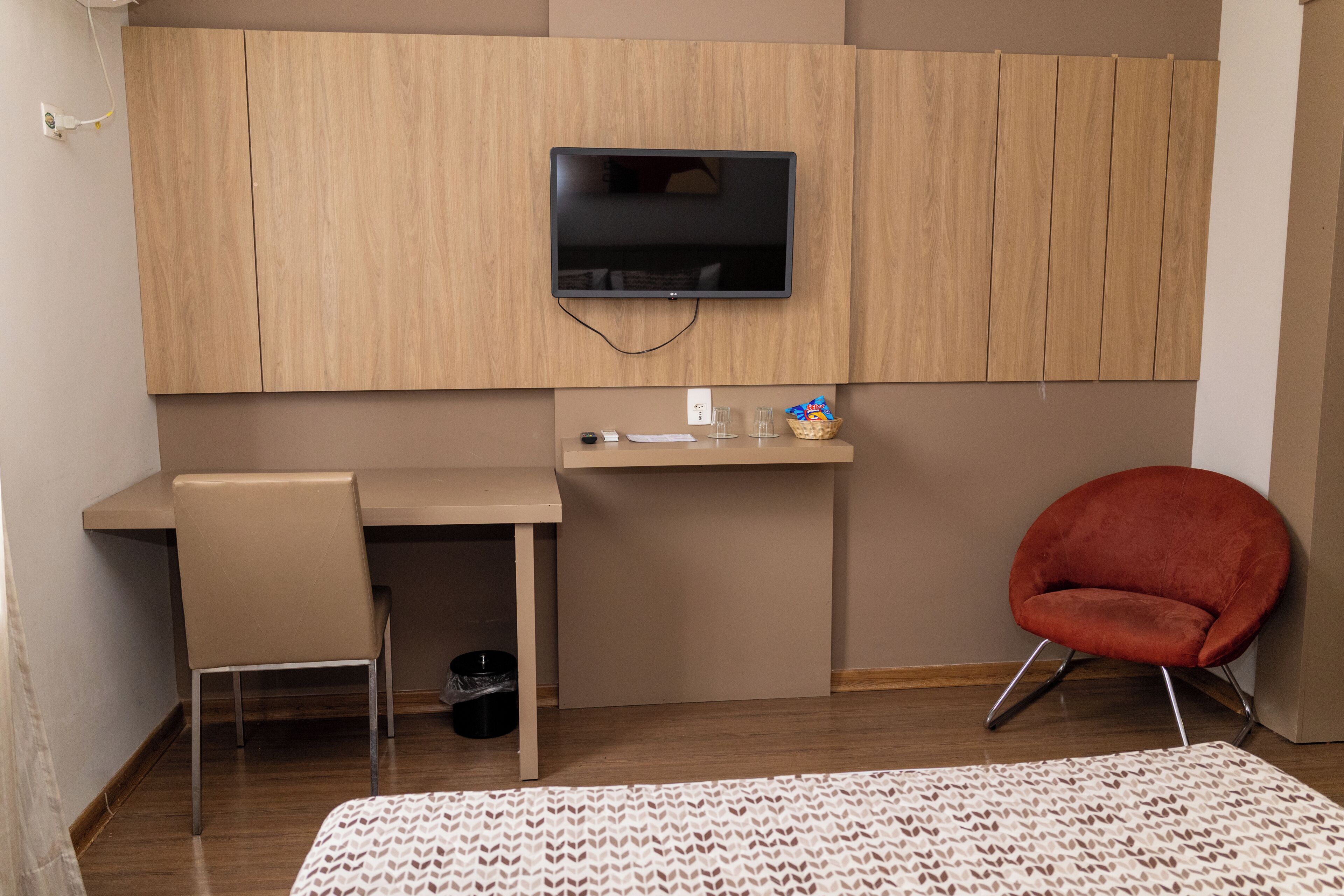 A modern room with a wooden wall, a flat-screen TV mounted above a desk, a red chair, and a patterned rug.