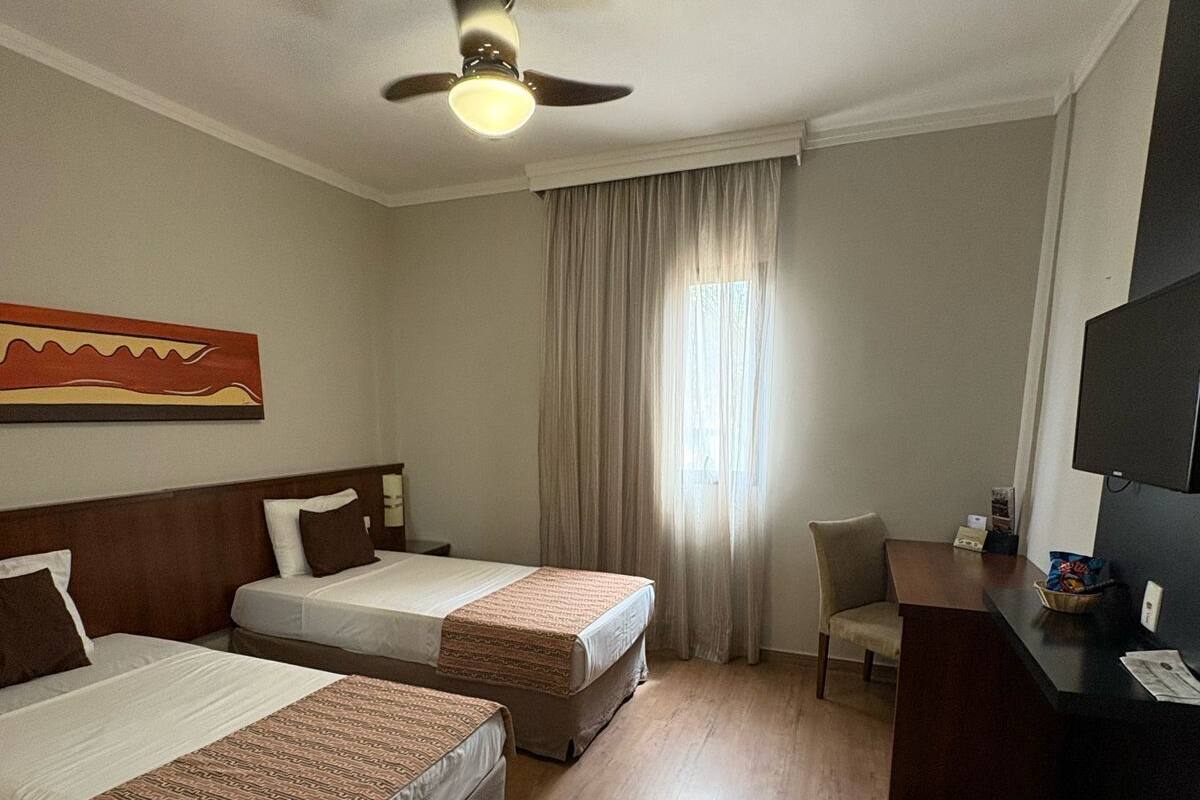 A hotel room with two beds, a desk, a chair, a ceiling fan, and a television.