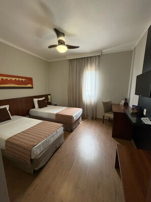 Ein Hotelzimmer mit zwei Betten, einem Schreibtisch, einem Stuhl, einem Deckenventilator und einem Fernseher.