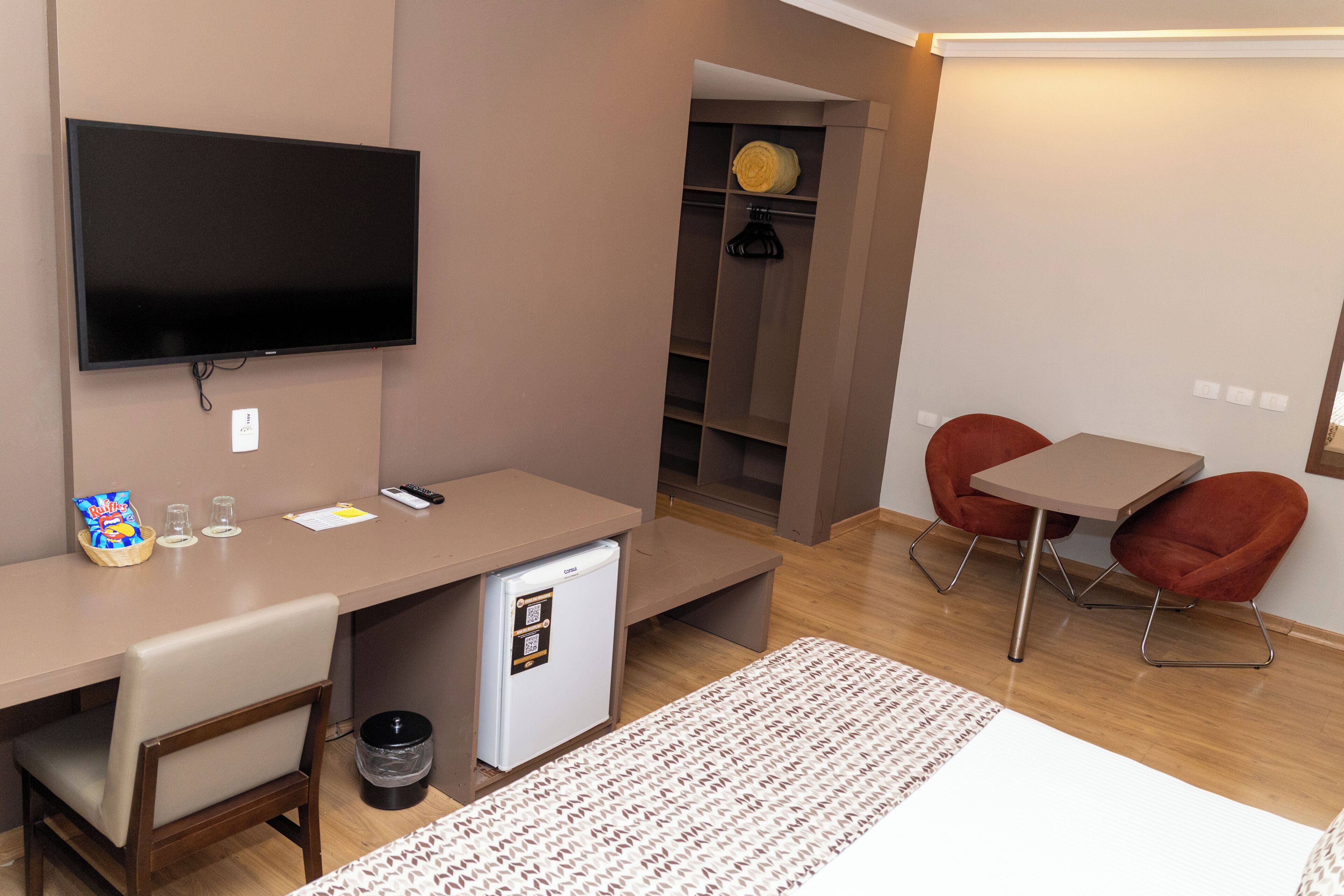 Habitación de hotel compacta con televisión de pantalla plana, refrigerador pequeño, escritorio, dos sillas y una cama.