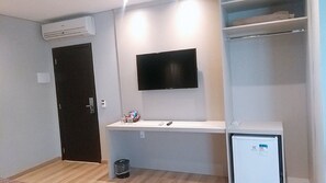 Une petite pièce moderne, avec un bureau blanc, un téléviseur à écran plat fixé au mur au-dessus, un petit réfrigérateur et une porte dotée d’un cadre foncé.