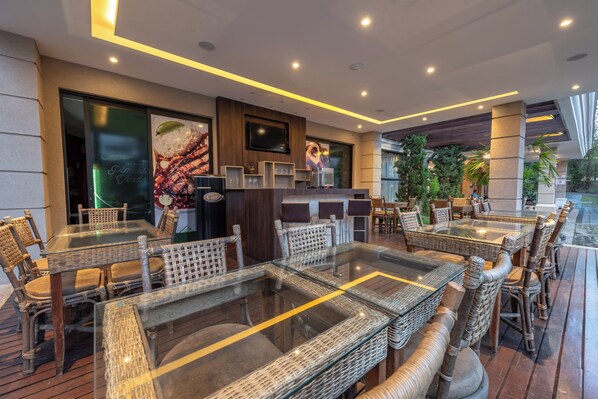 A restaurant with glass-top tables and wicker chairs, a flat-screen TV mounted on the wall, and a view of the outdoors.