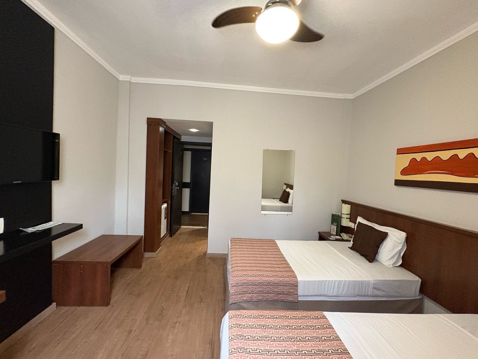 Een hotelkamer met twee bedden, een televisie, een plafondventilator, een spiegel en een schilderij aan de muur.