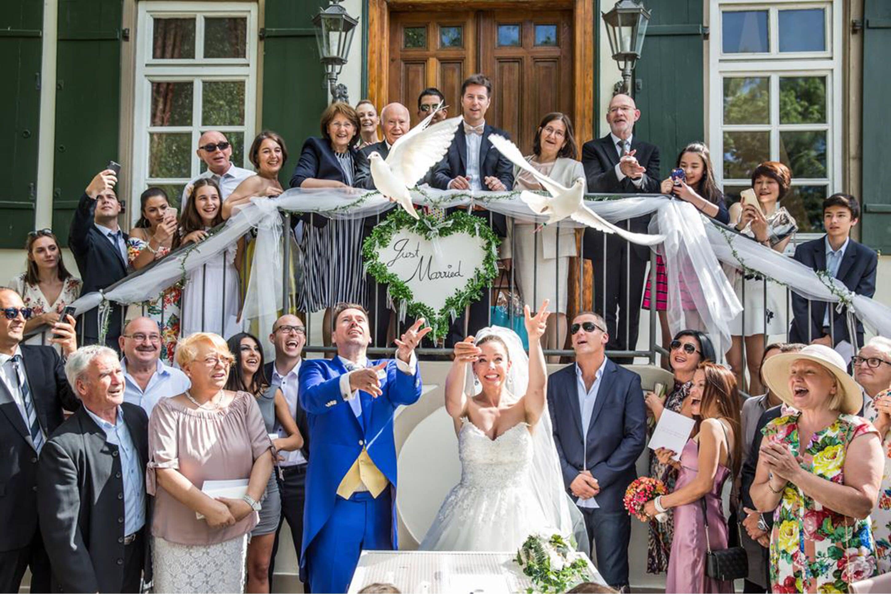 Un grupo de personas en una ceremonia de boda, con un cartel que dice 'Just Married' y palomas volando.