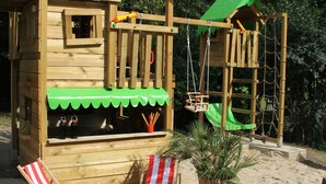 A wooden playhouse with a slide, climbing net, and a small bridge. There are two beach chairs and a potted plant nearby.