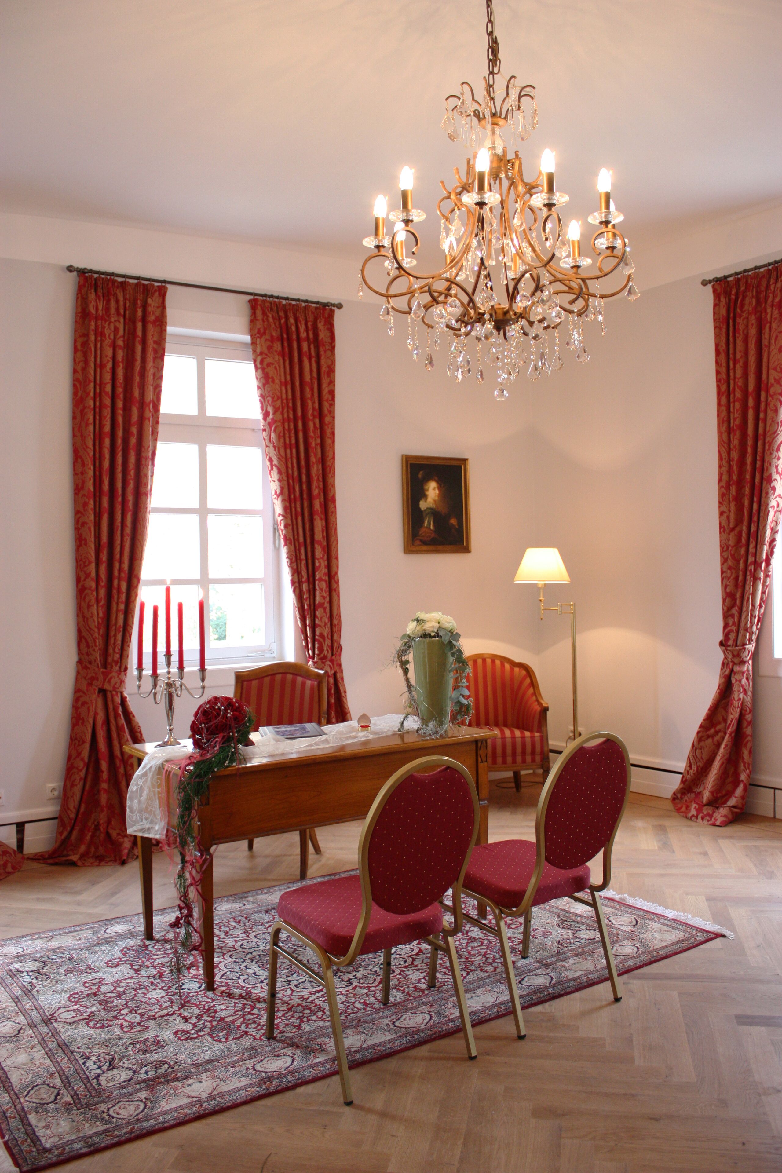 A dining room with a chandelier, red curtains, a table with chairs, a vase with flowers, and a lamp.