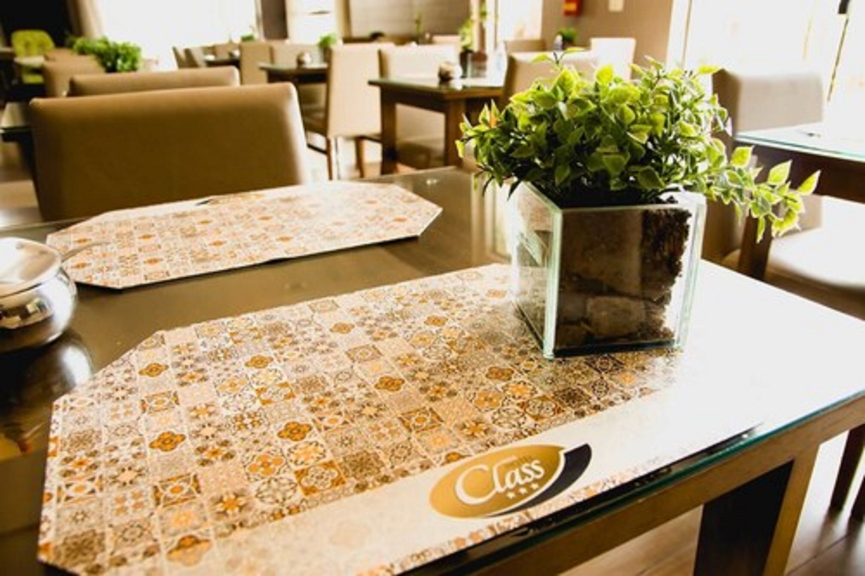 Una mesa de comedor con un mantel individual estampado y una jara de vidrio con una planta.