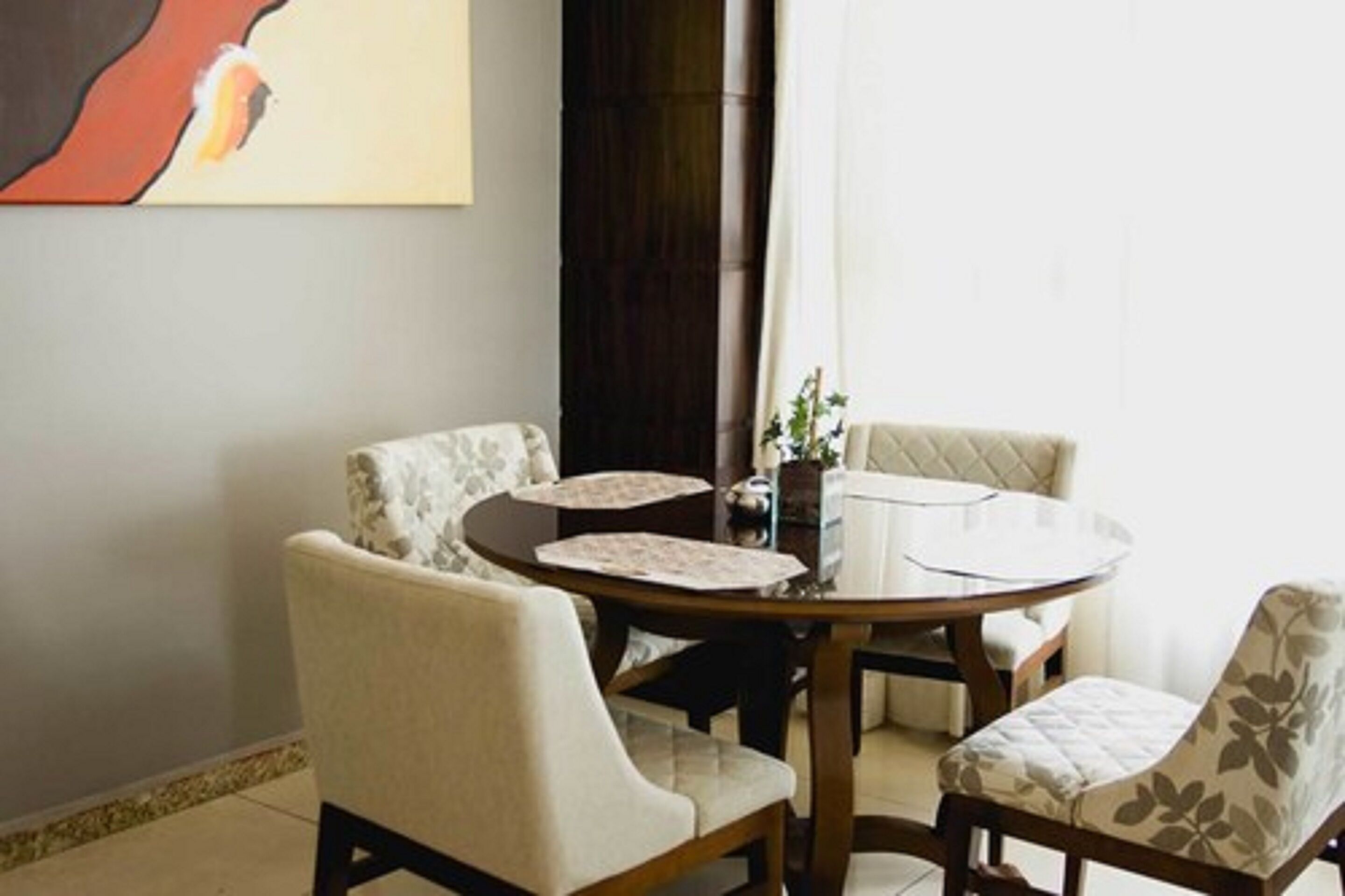 Een eetgedeelte met een ronde tafel en vier stoelen, een schilderij aan de muur en een donker gordijn.