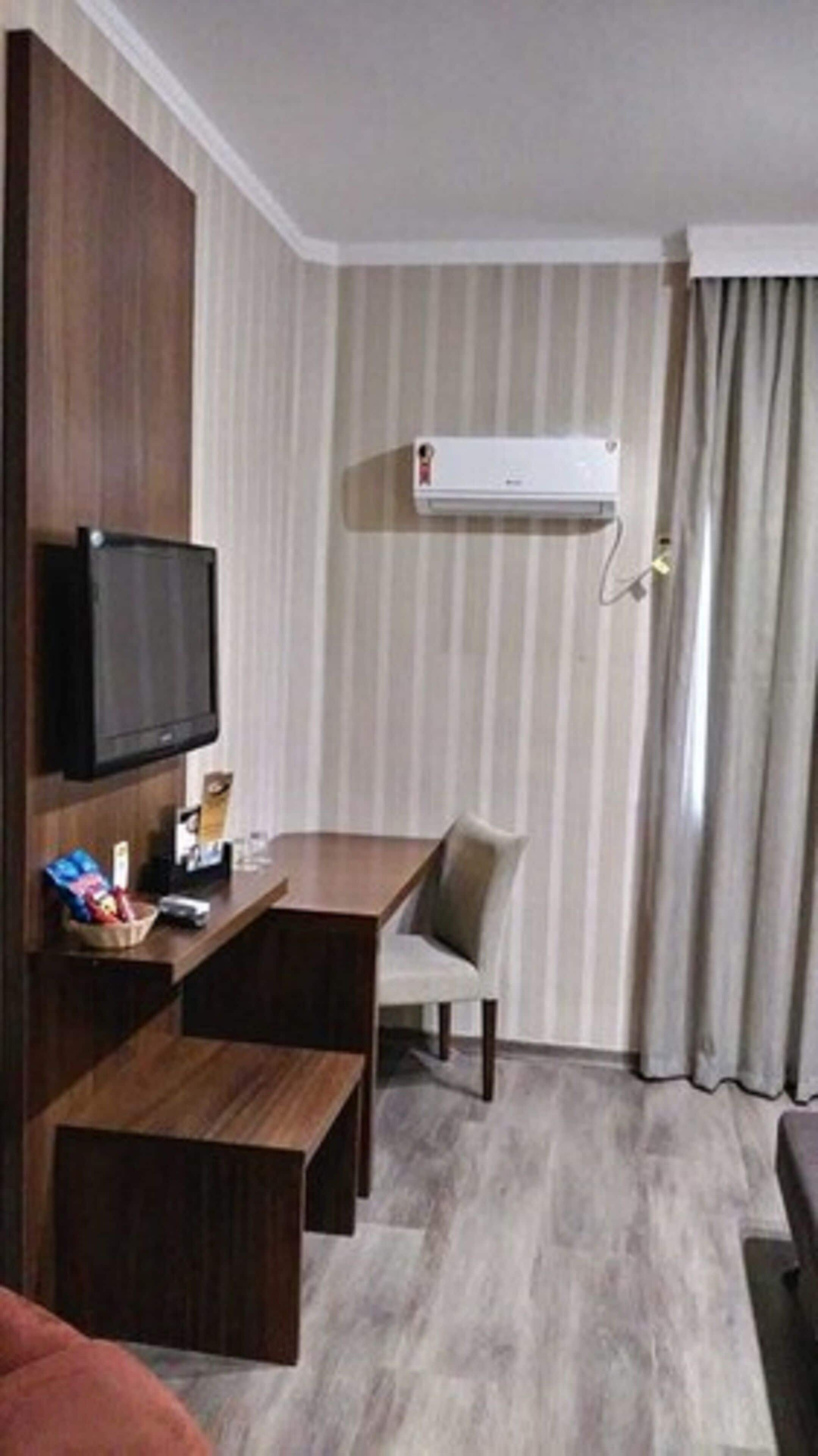 Habitación de hotel con escritorio de madera, televisor montado en la pared, ventana con cortinas y equipo de aire acondicionado.