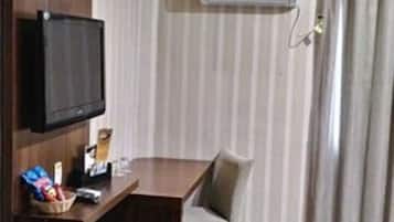 A hotel room with a wooden desk, a television mounted on the wall, a window with curtains, and an air conditioning unit.