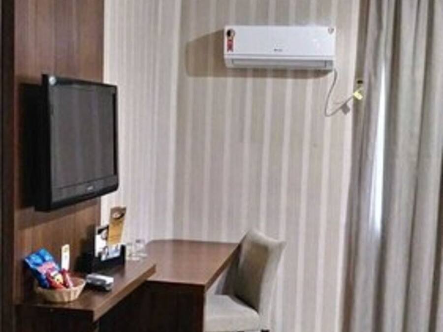 A hotel room with a wooden desk, a television mounted on the wall, a window with curtains, and an air conditioning unit.