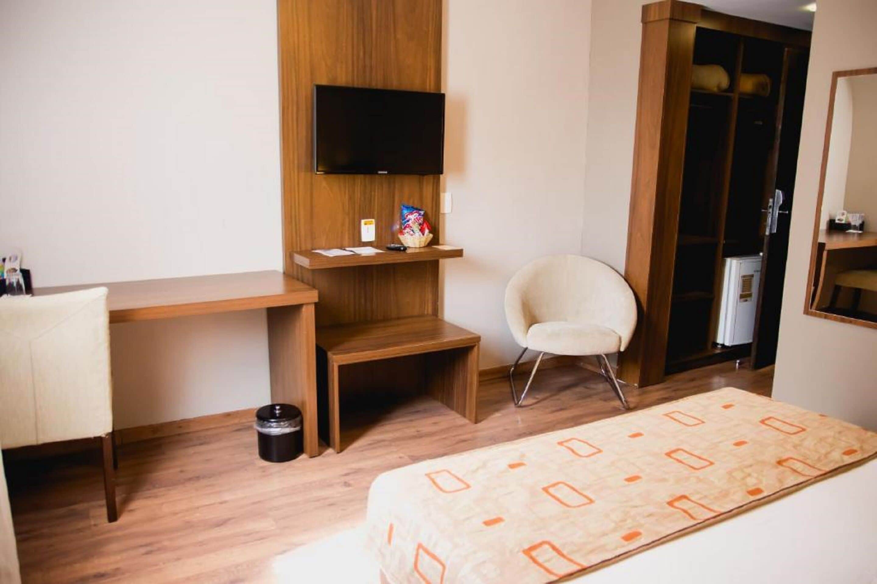 Une chambre d’hôtel avec un bureau en bois, une chaise, un lit recouvert d’une couvre-lit à motifs, un téléviseur fixé au mur et une armoire.