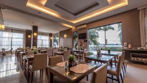 A modern hotel dining area with multiple tables and chairs, large windows with a view of palm trees, and a kitchen area visible in the background. - Class Hotel Varginha (Varginha)