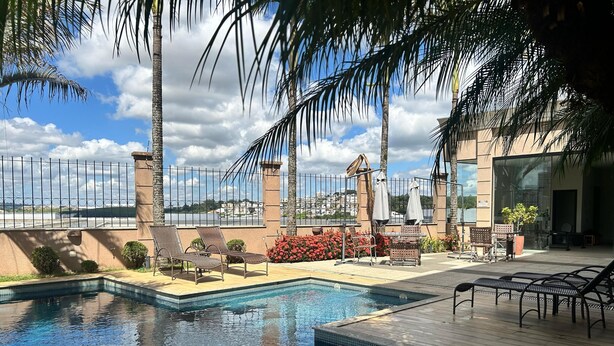 A swimming pool with lounge chairs and umbrellas, surrounded by palm trees and a view of a cityscape. - Class Hotel Varginha (Varginha)