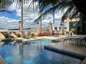 A swimming pool with lounge chairs and umbrellas, surrounded by palm trees and a view of a cityscape. - Class Hotel Varginha (Varginha)