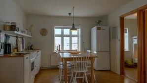 A kitchen with white cabinets, a wooden dining table, and chairs. There is a window with a view of the outside, a refrigerator, and a clock on the wall. - Gamla húsið - The old house (Sveitarfélagid Ölfus)