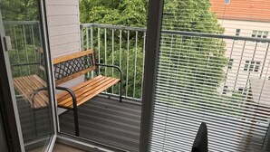 Ein Balkon mit Holzbank, Metallgeländer und Blick auf Grünflächen und Gebäude durch Jalousien.