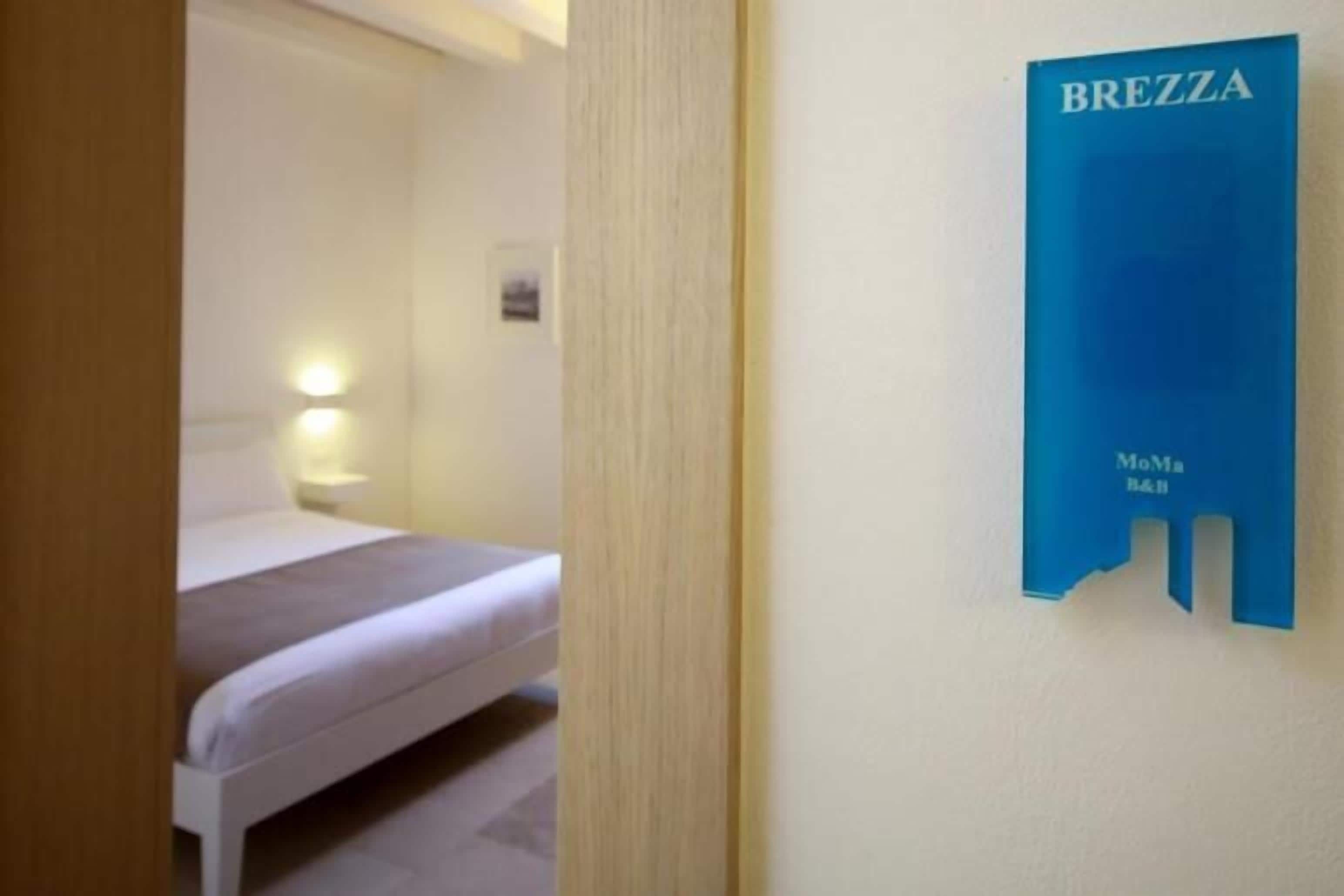 A hotel room with a bed, a nightstand with a lamp, and a blue sign on the wall that reads "BREZZA MoMa B&H".