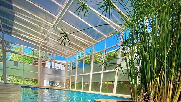 A swimming pool with clear blue water, surrounded by a covered area with large windows and tropical plants. - Brisas de Villa Gesell (Villa Gesell)