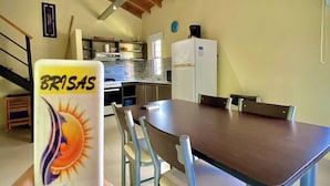 A dining area with a wooden table and chairs, a kitchen in the background, and a hand holding a card with a sun design. - Brisas de Villa Gesell (Villa Gesell)
