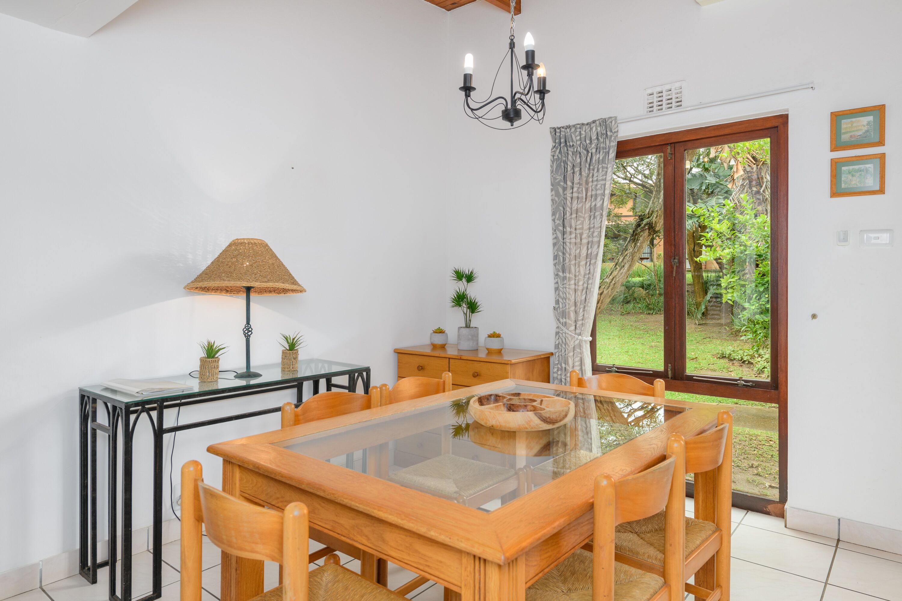 A dining area with a glass-top table, wooden chairs, a chandelier, a lamp, and a view of the garden through glass doors.