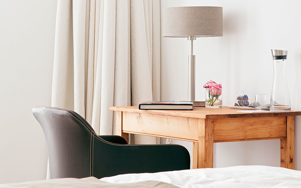 Habitación de hotel ordenada con un escritorio de madera, una silla, una lámpara, un jarrón con una flor y una botella de vidrio.