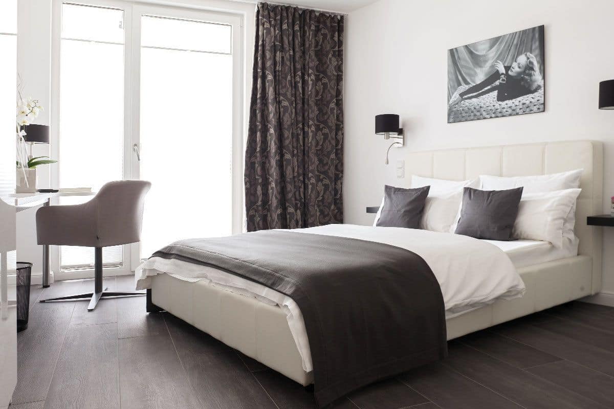 A modern bedroom with a large bed, a desk, and a chair. There is a window with curtains, a wall-mounted lamp, and a picture on the wall.