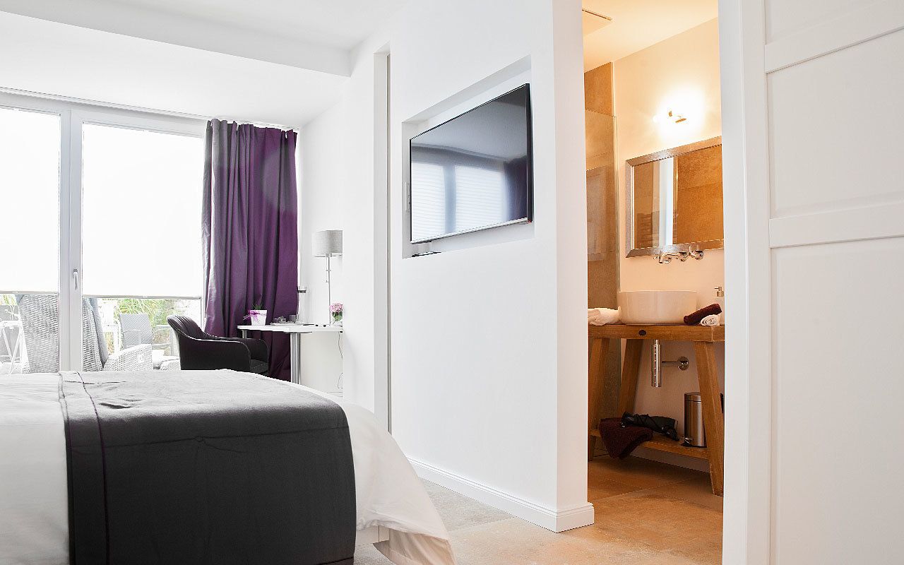 A hotel room with a bed, a desk with a chair, a TV mounted on the wall, a bathroom with a sink and mirror, and a balcony with a view.