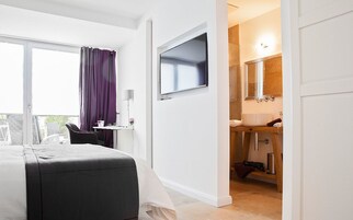 Habitación de hotel con cama, escritorio con silla, televisor montado en la pared, baño con lavamanos y espejo, y balcón con vistas.