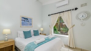 A neatly made bed with a blue and white color scheme, a painting of a beach scene, a wall clock, and a window with curtains. - San Lameer Villa Rentals  2301 (Southbroom)