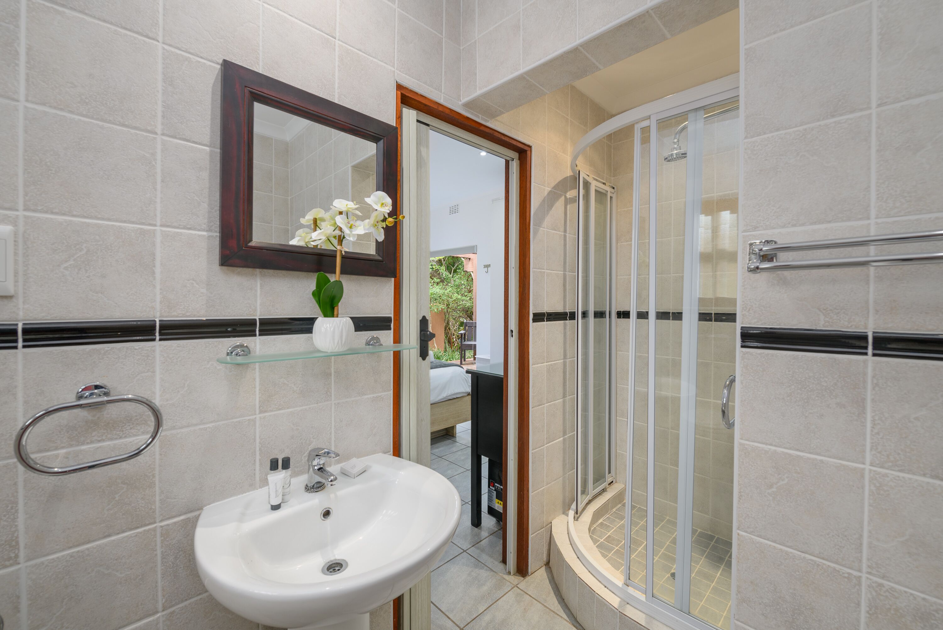 Une salle de bain avec un lavabo blanc, une cabine de douche en verre et un miroir orné d’une orchidée blanche.