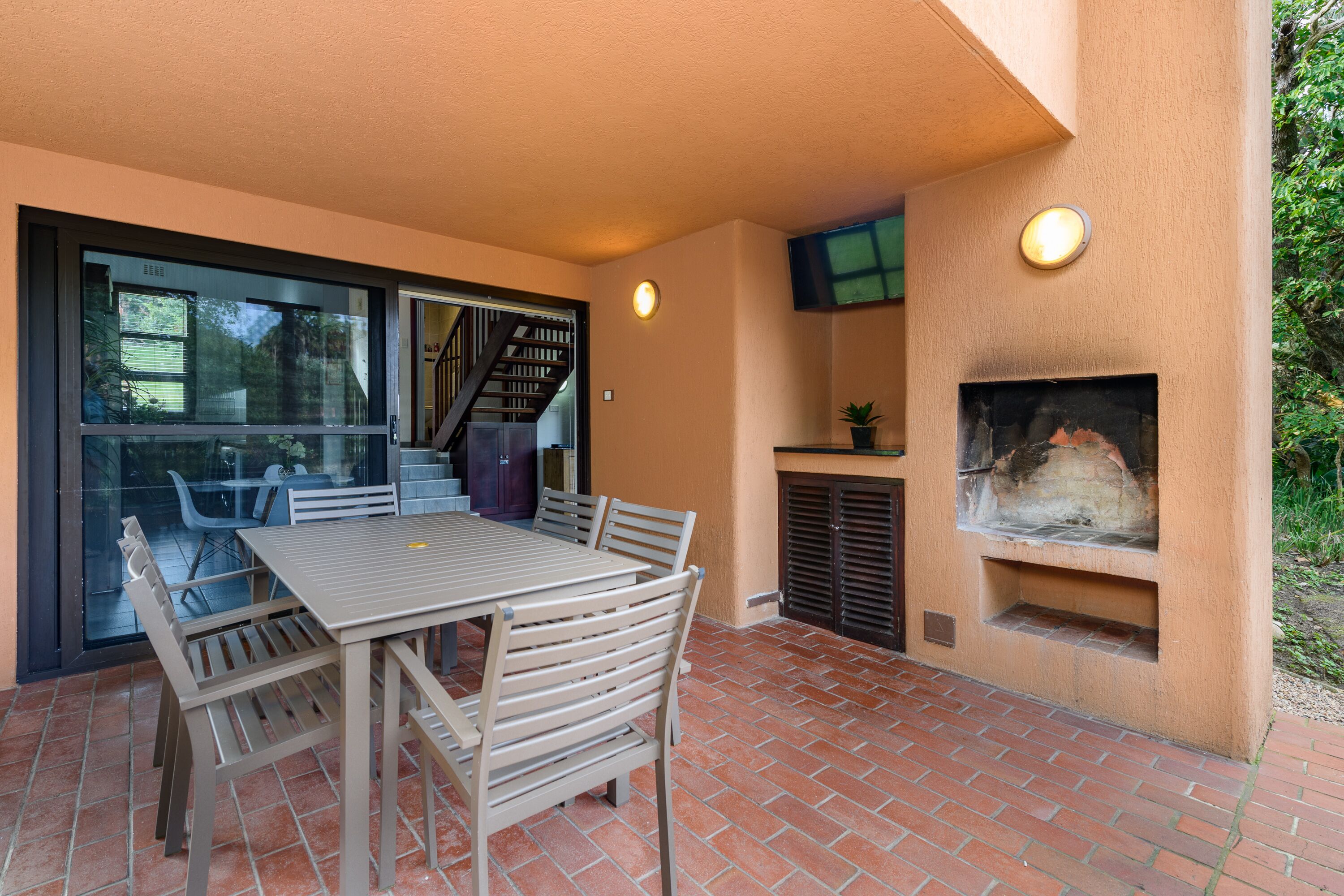 Un patio al aire libre con piso de ladrillo, una mesa con sillas, un vidrio que da a una escalera y una parrilla empotrada.
