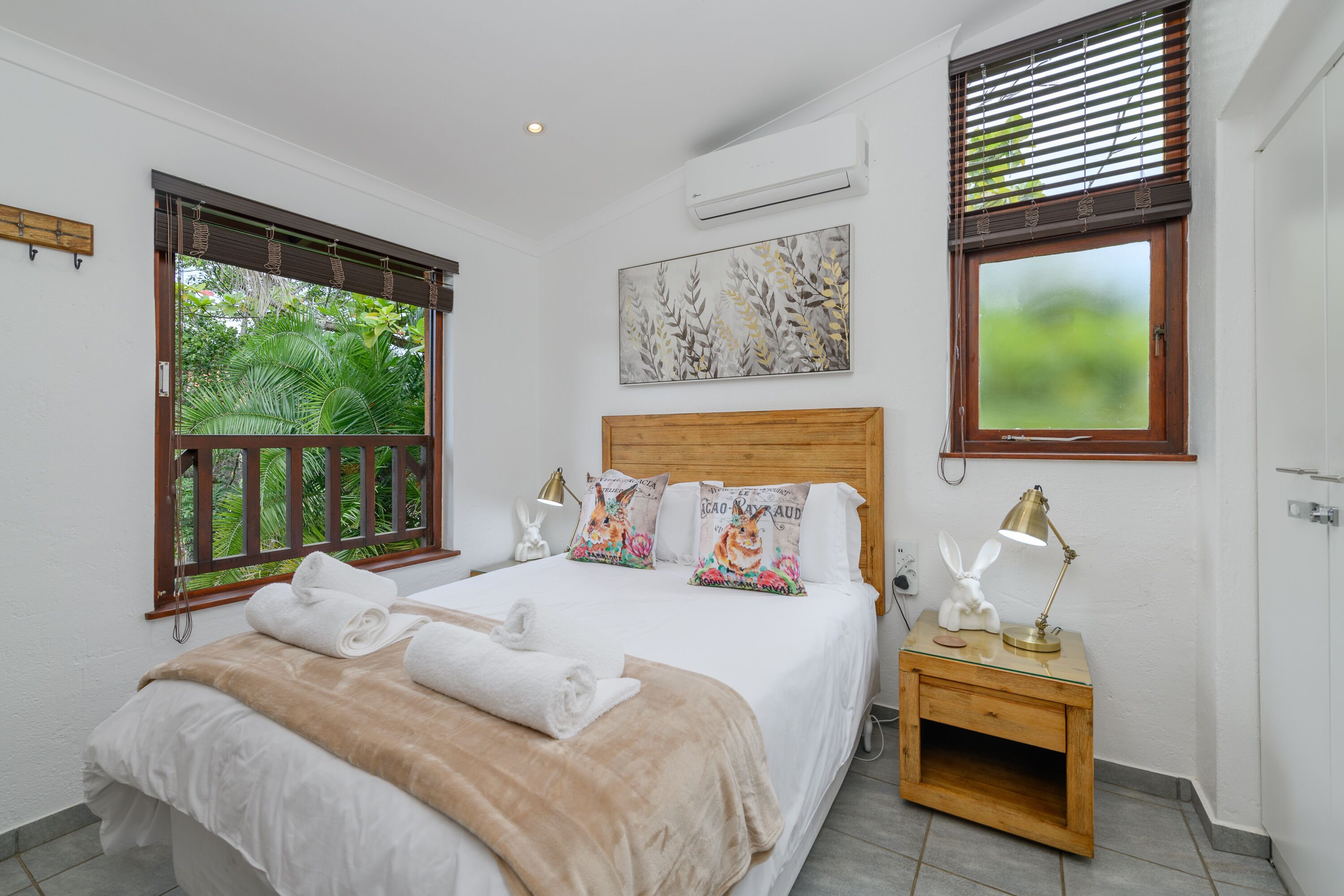 A neatly made bed with a wooden headboard, a bedside table with a lamp, and a window with blinds.