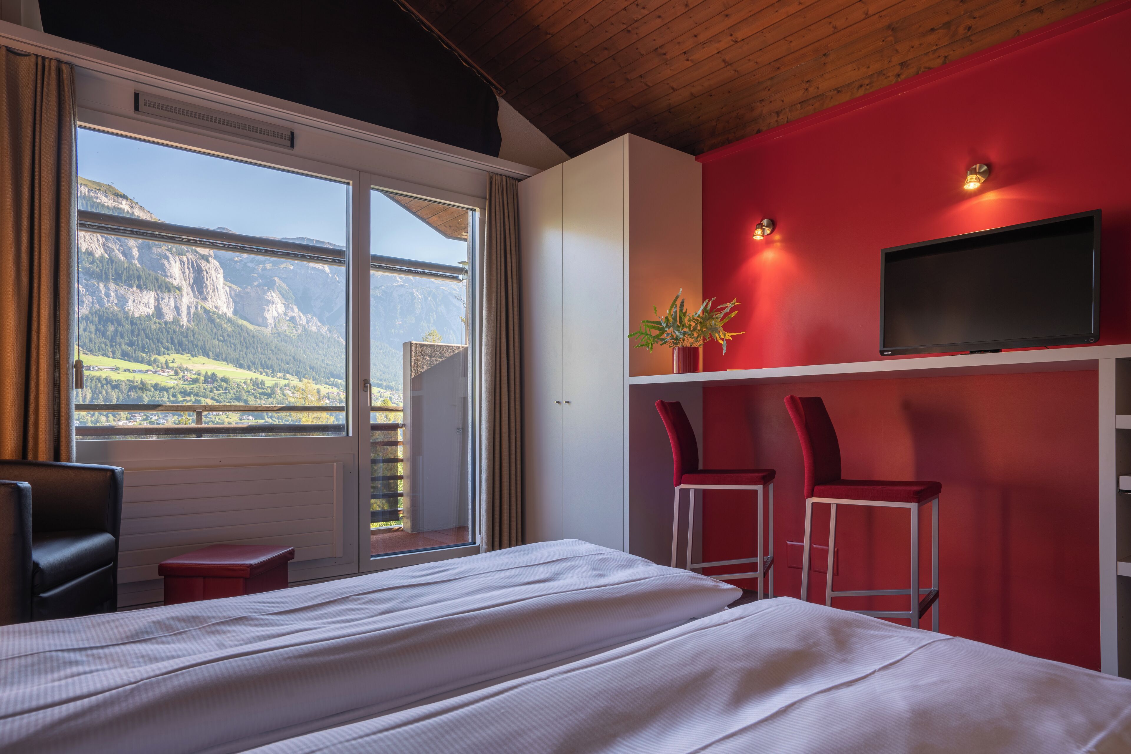 Una habitación de hotel con una cama, un televisor en una pared roja, dos sillas rojas, un ventanal con vista a la montaña y un banco rojo.