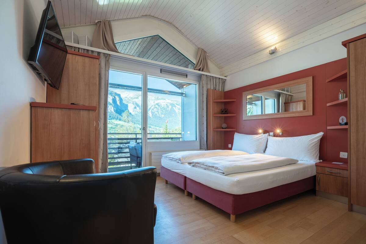 A bedroom with a bed, a TV mounted on the wall, a window with a view of mountains, and a chair.