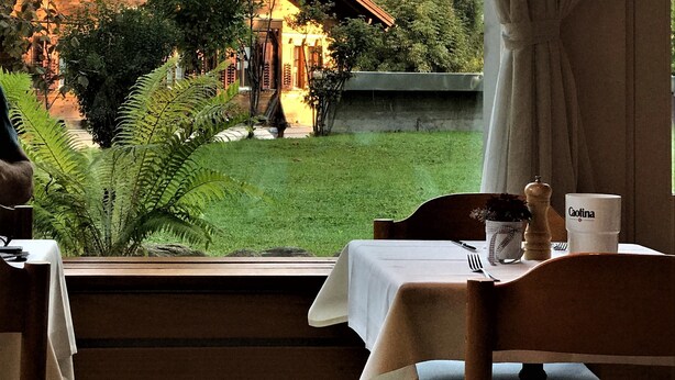 Uma sala de jantar com mesa posta para dois, vista de uma montanha e um edifício rústico ao fundo.