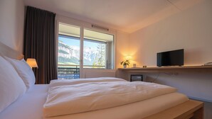 Quarto de hotel com uma cama grande, televisão em uma estante, janela com vista para as montanhas e abajur na mesa de cabeceira.