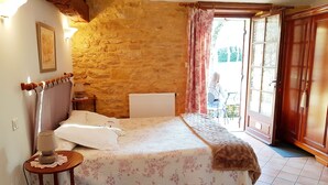 A bedroom with a stone wall, a bed with floral bedding, a bedside table with a lamp, and a view through an open door to the outside. - Les Filolies (Saint-Andre-d'Allas)