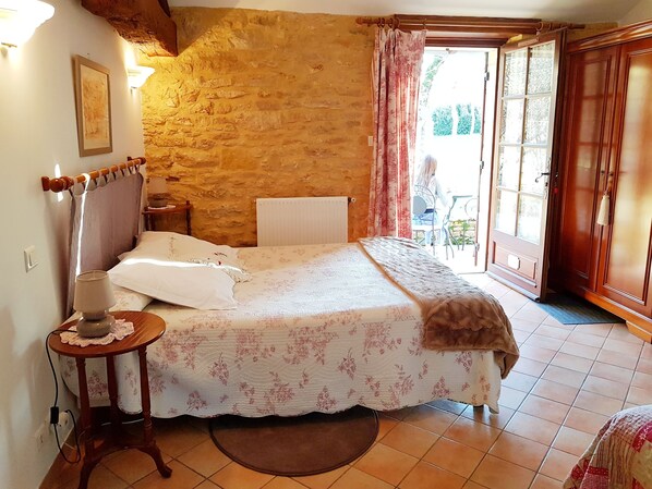 Une chambre avec un mur en pierre, un lit recouvert d’une literie à motifs floraux, une table de chevet avec une lampe, et une vue sur l’extérieur par une porte ouverte.