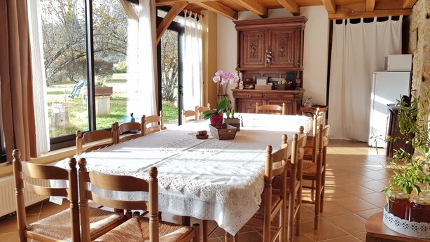 A dining area with a wooden table and chairs, a lace tablecloth, and a view of the outdoors through large windows. - Les Filolies (Saint-Andre-d'Allas)